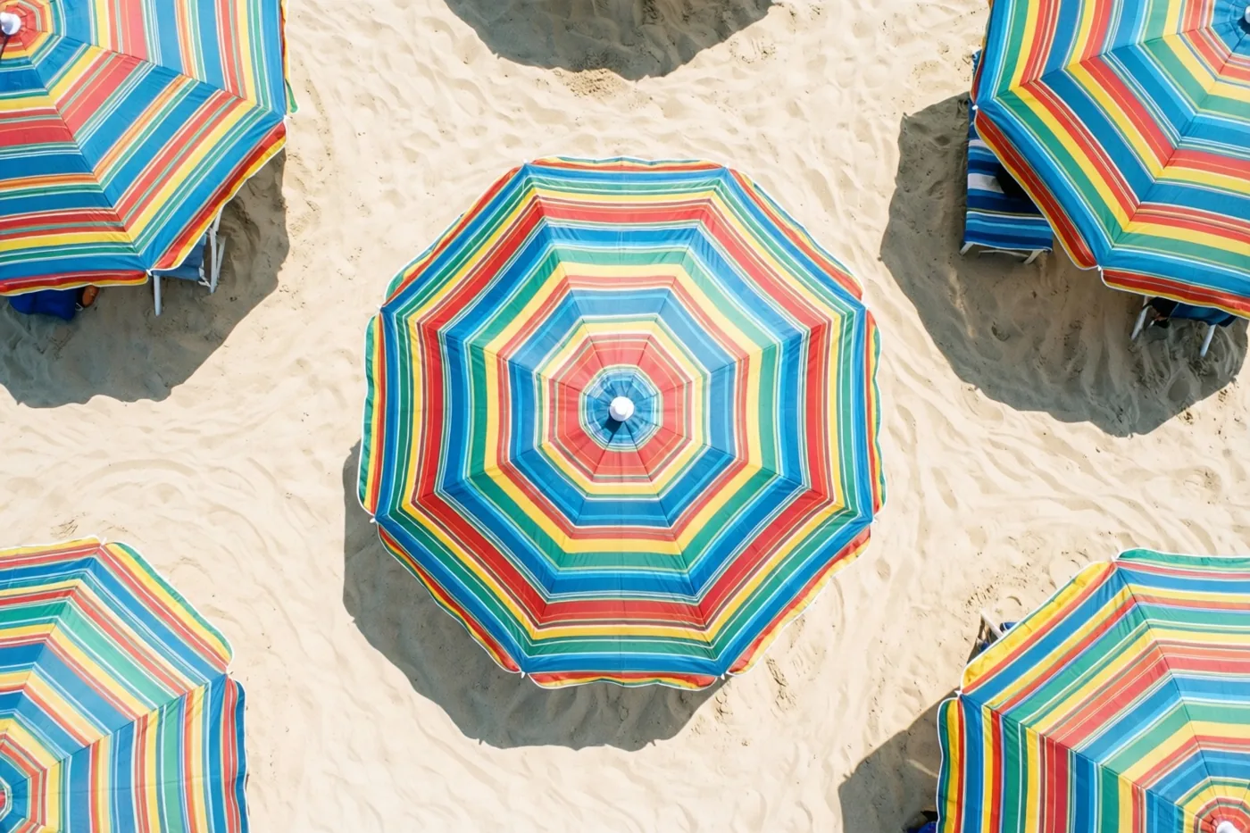 Verschillende strandparasols met blauwe, rode, gele en groene strepen staan op licht zand, gezien van bovenaf.