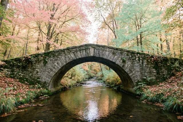 Een oude stenen voetbrug met één boog overspant een rustige beek in een herfstbos, in zachtroze, groene en aardbruine tinten.