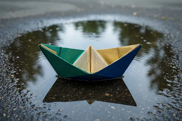 Een bootje gevouwen uit een boekpagina drijft op een plas in een omgeving die op een speelplaats lijkt, in groene, blauwe en gele tinten.