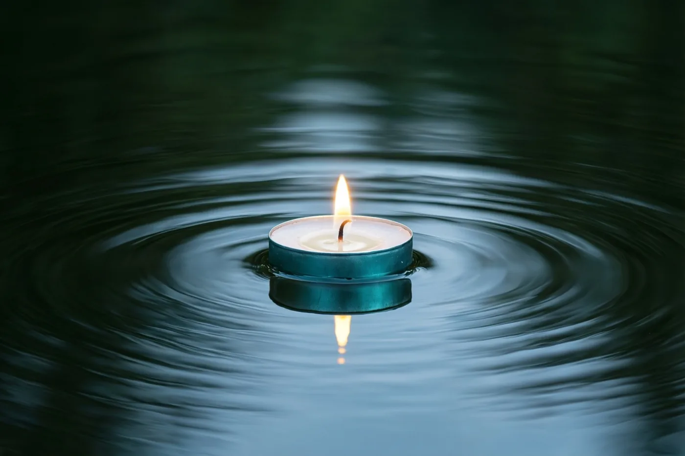 Een brandend theelichtje drijft op donker, stil water met kringen eromheen, in diepe blauwe en blauwgroene tinten.