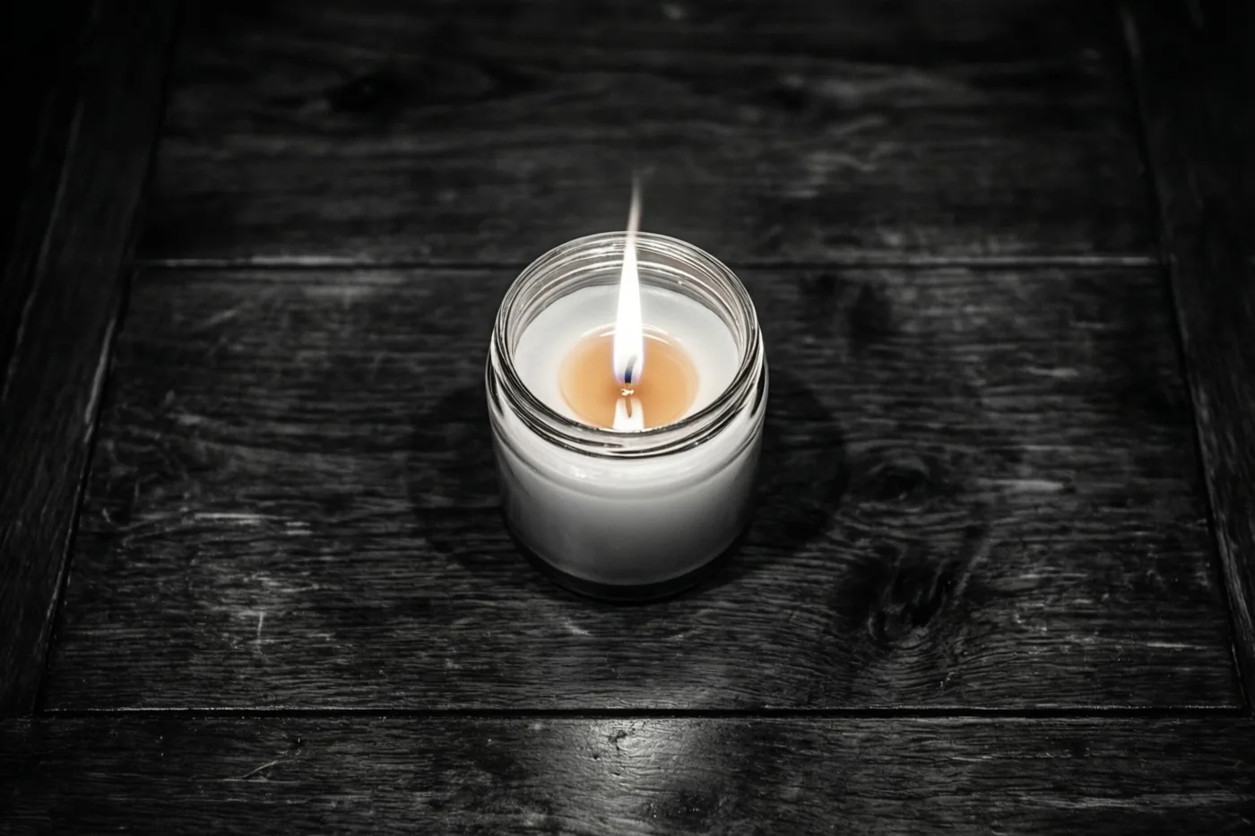 Een kaars in een glazen pot brandt op een donkere houten tafel, met een warm oplichtende vlam in een verder monochroom beeld.