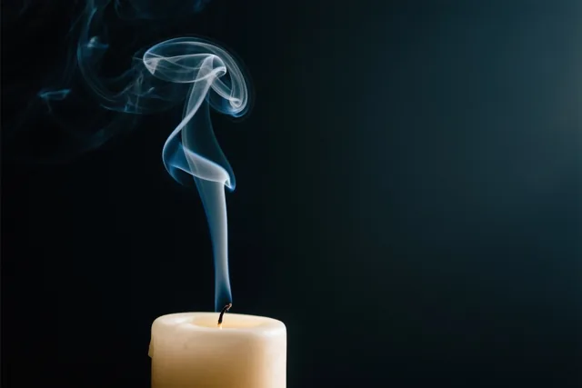 Rook krult omhoog uit een brandende kaars tegen een donkere achtergrond in zwarte, witte en crèmekleurige tinten.
