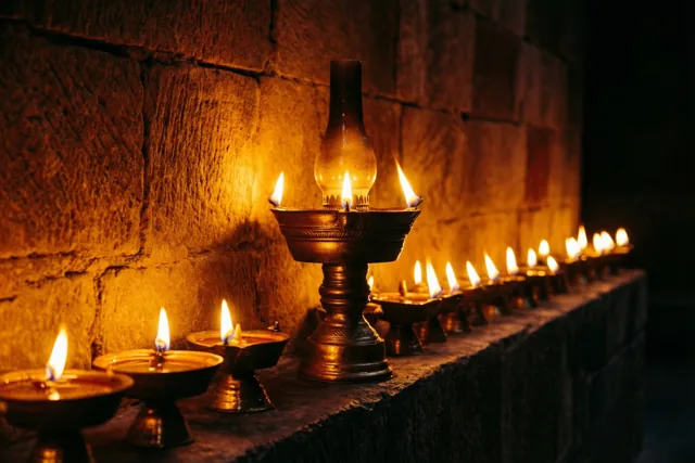 Brandende olielampen met een hogere middelste lamp staan op een stenen richel tegen een ruwe stenen muur, met gouden licht en diepe schaduwen.