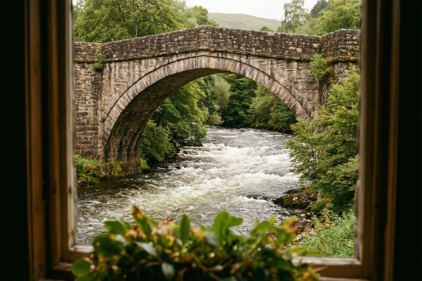 Door een houten raamkozijn is een stenen boogbrug over een snelstromende rivier te zien, met bos en heuvels erachter.