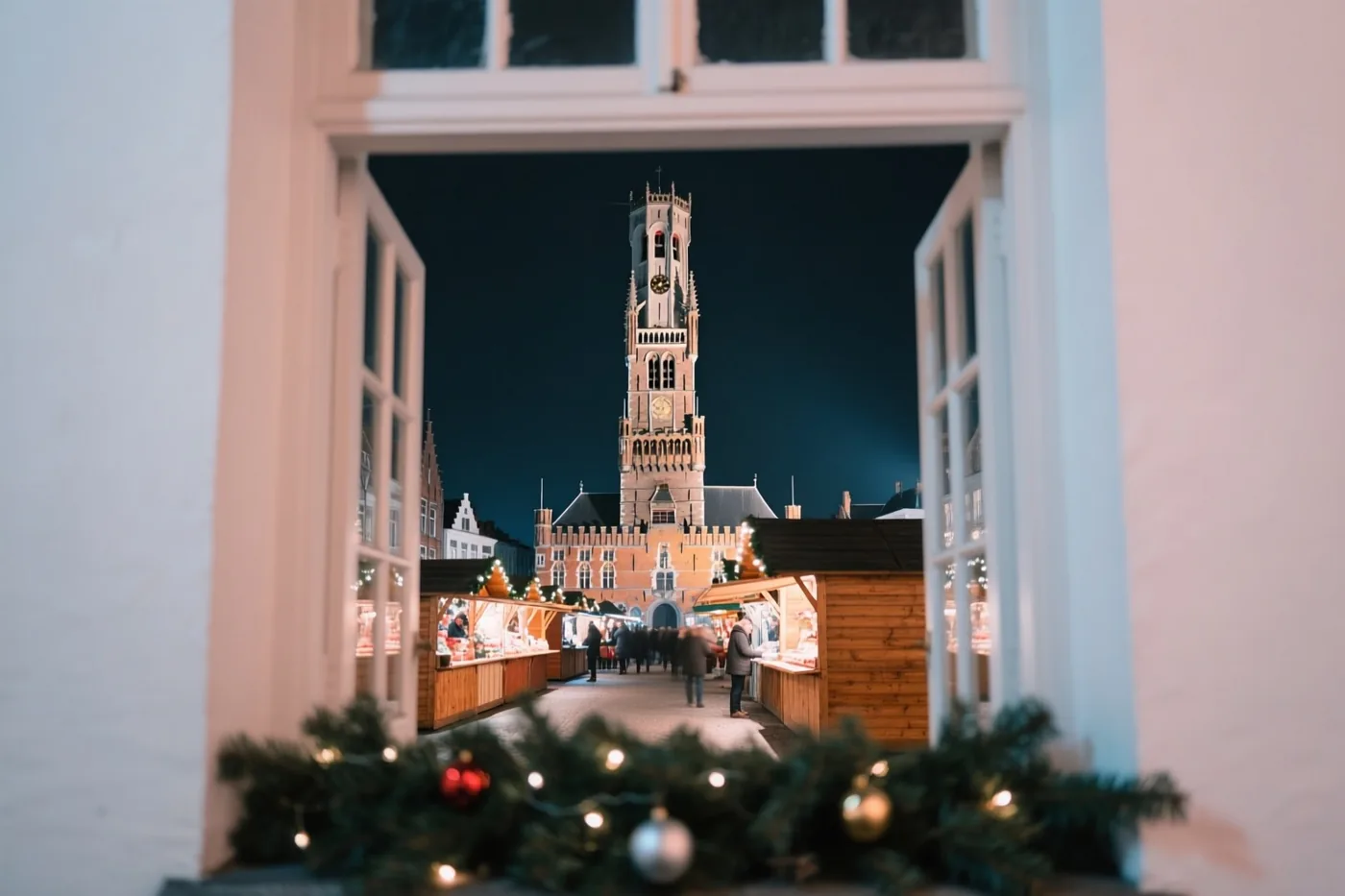 Door een open raam met groen en lichtjes op de vensterbank is een nachtelijke kerstmarkt in Brugge te zien, met houten kraampjes en het verlichte Belfort.
