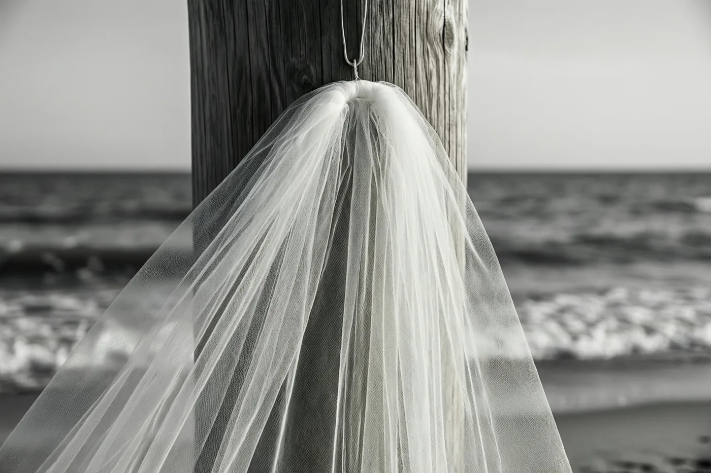 Een zwart-witfoto toont een witte bruidssluier aan een verweerde houten strandpaal, met de zee en golven op de achtergrond.
