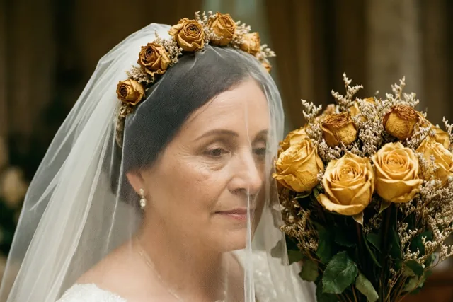 Een detailopname toont een vrouw met een bruidssluier versierd met gedroogde goudkleurige rozen, die een gedroogd bruidsboeket vasthoudt naast een vers boeket goudgele rozen.