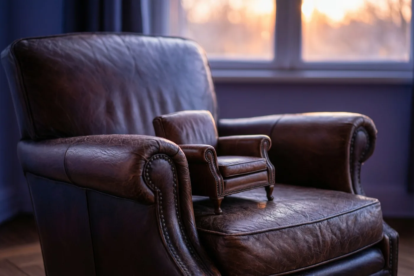 Een grote bruine leren fauteuil en een kleine bijpassende fauteuil staan naast elkaar in een kamer met warm zonlicht door het raam.