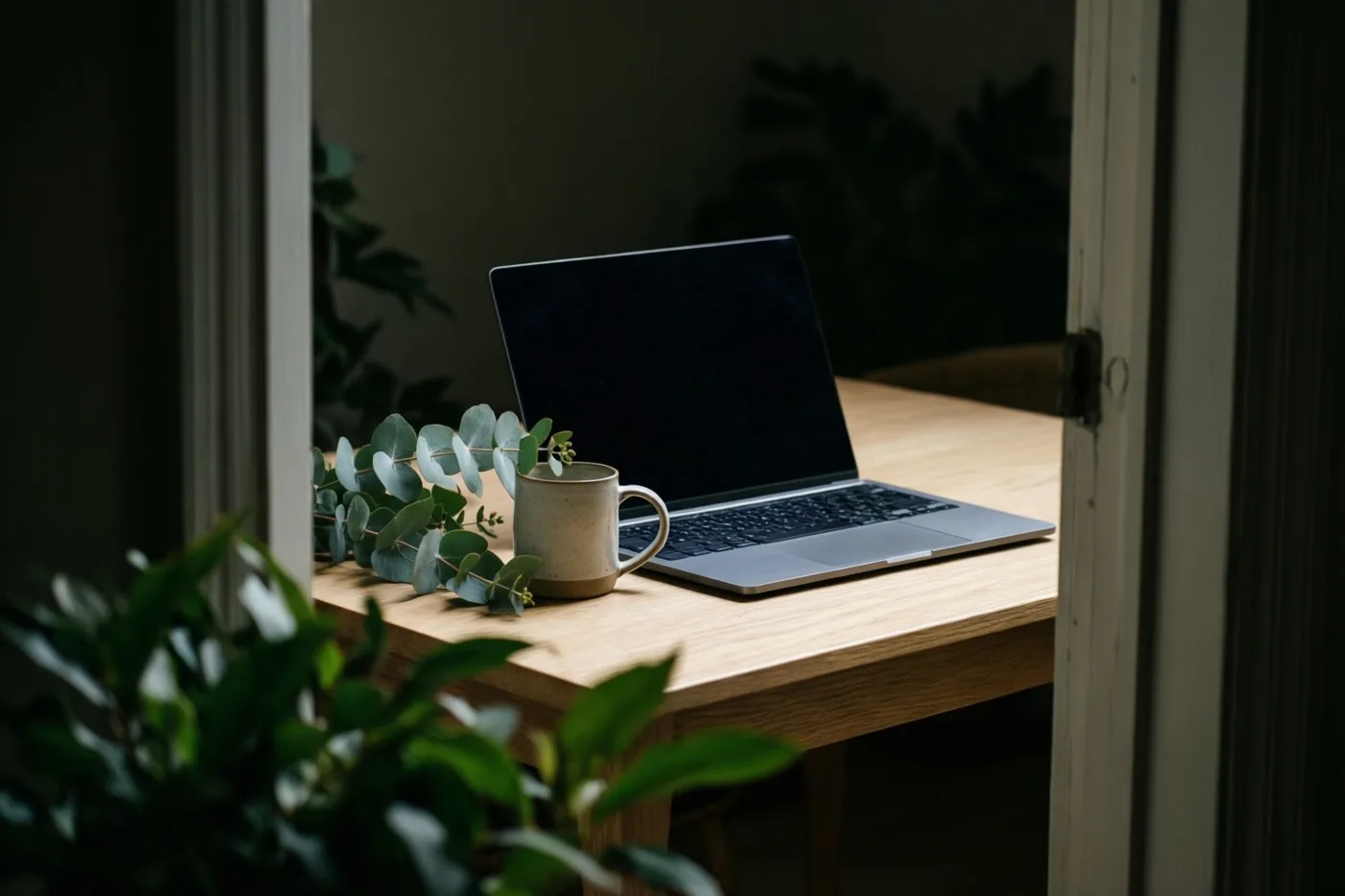 Door een deuropening zijn een zilverkleurige laptop, een beige keramieken mok en een takje eucalyptus op een licht eiken bureau te zien, in zacht daglicht.