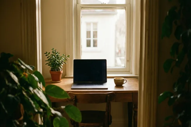 Een houten bureau bij een raam draagt een laptop, een klein potplantje en een kop koffie in warm daglicht.