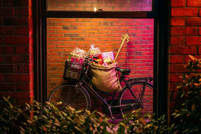Door een raam is een fiets met een jute zak en ingepakte cadeaus tegen een bakstenen muur te zien, in warm rood en roze licht.