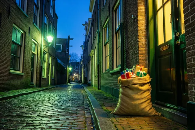Een jute zak vol kleurrijke pakjes staat op de drempel van een bakstenen huis in een smal kasseiensteegje, onder warme straatlampen en een diepblauwe avondhemel.