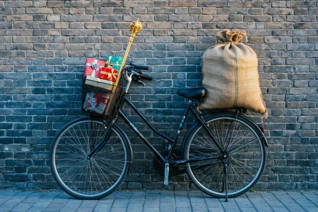 Een zwarte fiets staat in warm zonlicht tegen een donkere bakstenen muur, met een jute cadeauzak op de bagagedrager en ingepakte cadeaus en een gouden staf in de voormand.