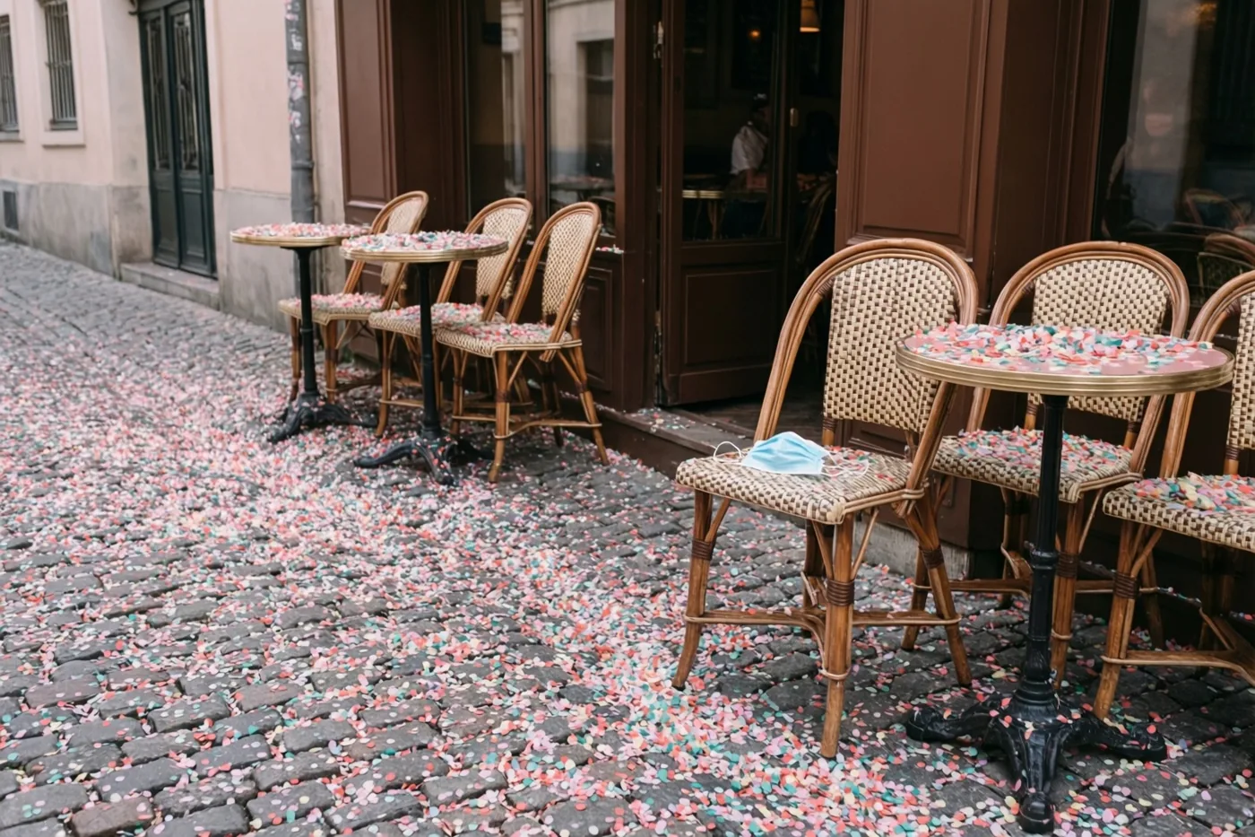 Kleurrijke confetti bedekt de kasseien voor een bruin café met buitentafels en stoelen, waarvan één een blauw mondmasker draagt.