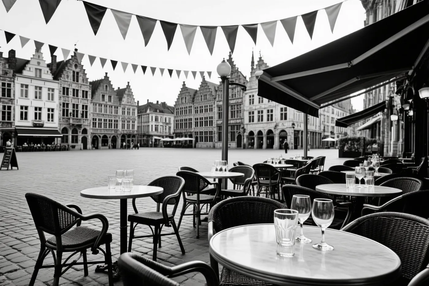 Een zwart-witfoto toont een leeg caféterras met tafels en stoelen onder vlaggetjesslingers op een kasseiplein, met historische gevels op de achtergrond.