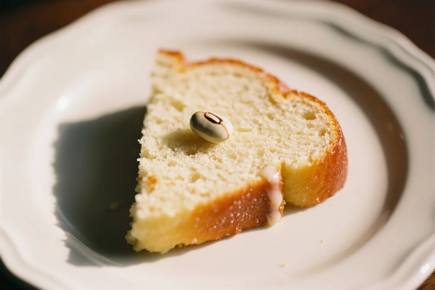 Een plak lichtgekleurde cake met een kleine ronde boon met een patroon erop ligt op een wit bord met een geschulpte rand in warm licht.