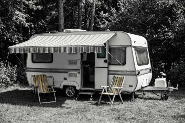 Een zwart-witbeeld toont een retrocaravan met een gestreepte luifel en twee bijpassende gestreepte klapstoelen op gras tussen de bomen.