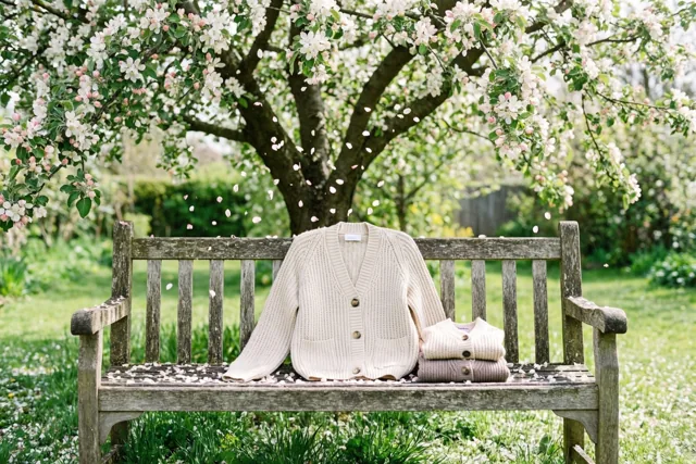 Een crèmekleurige cardigan en een kleinere, opgevouwen cardigan in gedempte kleuren liggen op een verweerde houten bank onder een bloeiende appelboom, met vallende bloemblaadjes in een zonnige tuin.