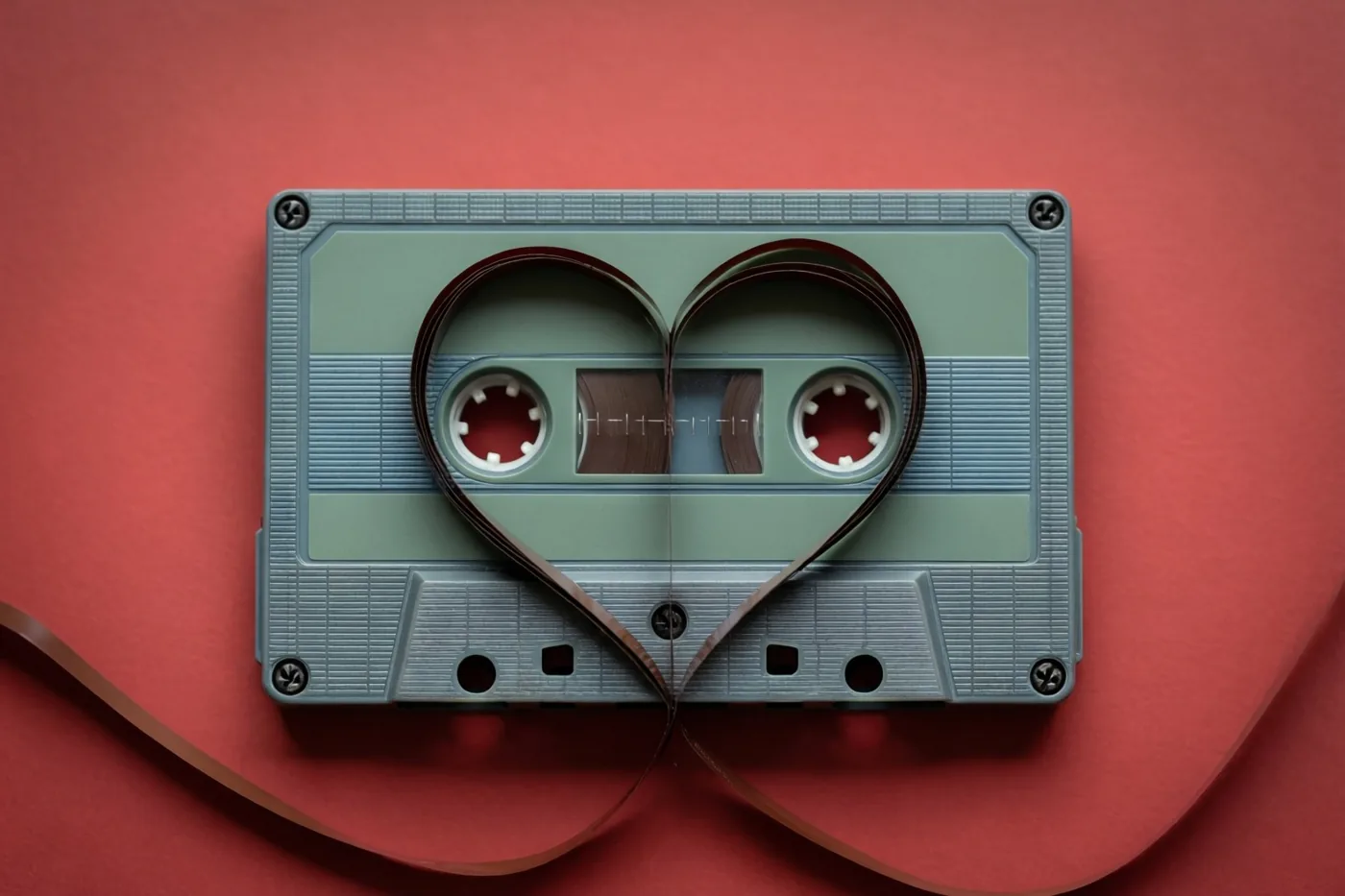 Een cassette met uitgetrokken magneetband in hartvorm ligt tegen een effen rode achtergrond, met blauwgroene en bruine tinten in het beeld.