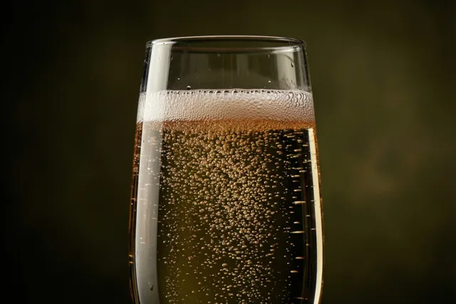 Een hoog glas champagne met opstijgende belletjes staat tegen een donkere, onscherpe achtergrond in goudbruine tinten.