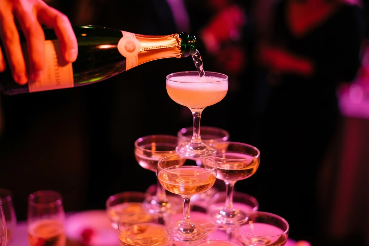 Een hand schenkt champagne in het bovenste glas van een piramide van champagnecoupes in een donkere feestruimte met warmroze en oranje licht.