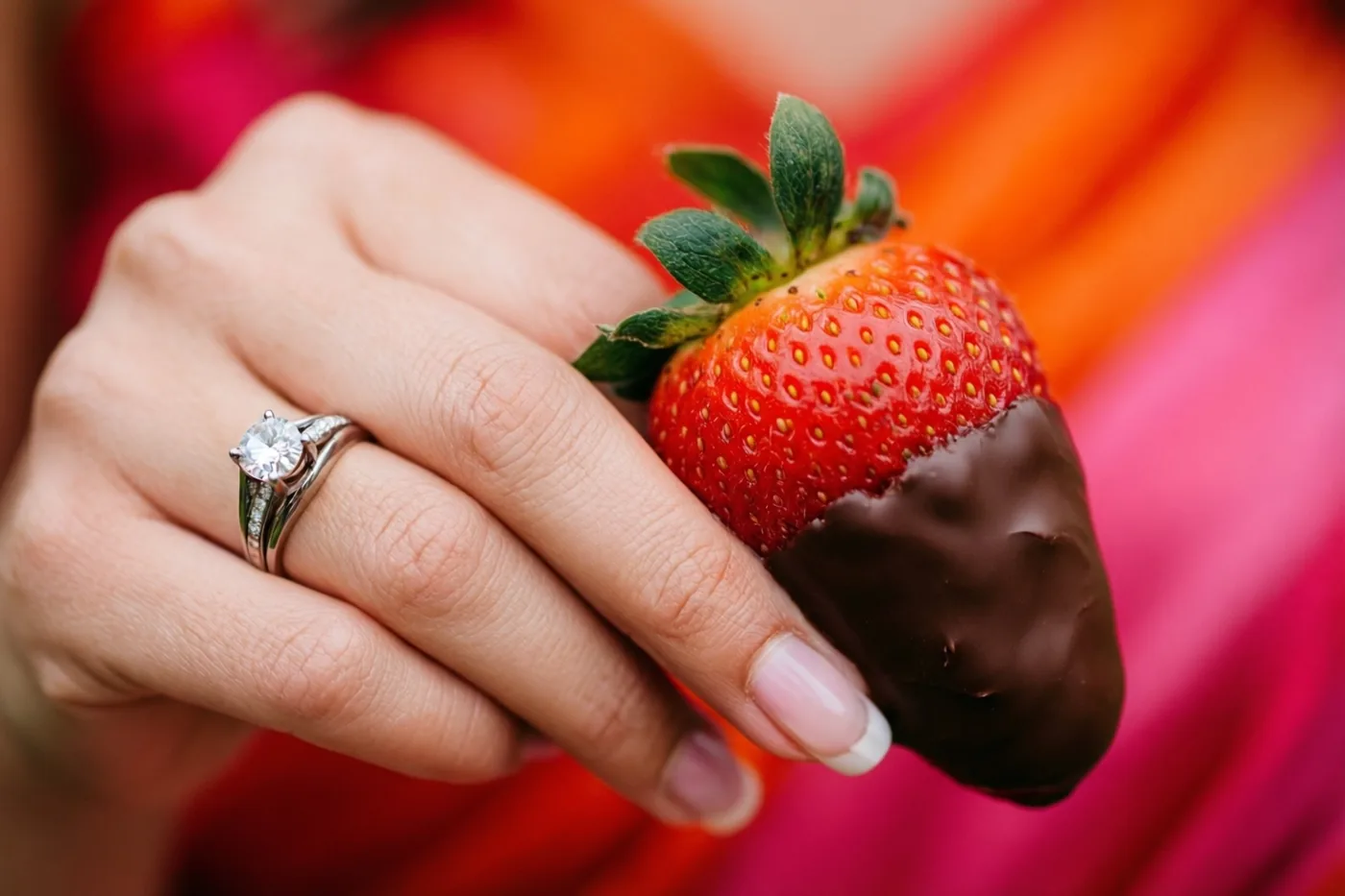 Een hand met een diamanten verlovingsring houdt een in chocolade gedoopte aardbei vast tegen een wazige achtergrond met rode, roze en oranje tinten.