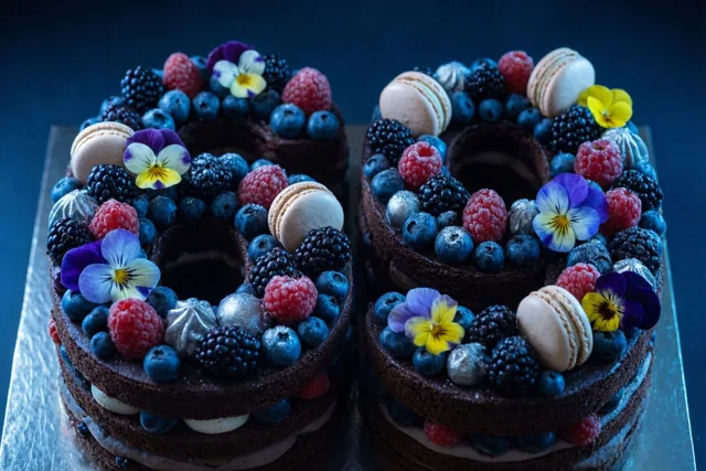 Twee chocoladetaarten vormen samen 69 en zijn versierd met frambozen, bramen, blauwe bessen, macarons en eetbare bloemen op een metalen ondergrond tegen een donkerblauwe achtergrond.