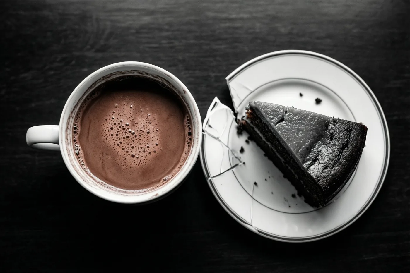 Van bovenaf zijn een witte mok warme chocolademelk en een stuk donkere cake op een gebarsten wit bord te zien op een donker houten oppervlak.