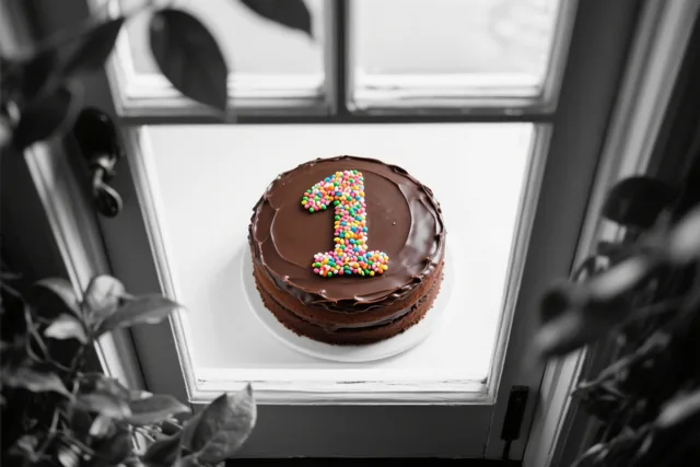 Een ronde chocoladetaart met het cijfer 1 van gekleurde suikerstrooisels staat op een wit bord op de vensterbank, omlijst door wazige bladeren in zwart-wit.