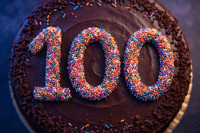 Een ronde chocoladetaart is van bovenaf te zien met 100 in gekleurde suikerstrooisels, donkerbruin glazuur en chocoladekruimels langs de rand.