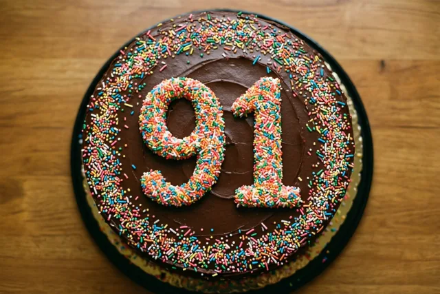 Van bovenaf is een ronde chocoladetaart op hout te zien, met 91 en een rijk versierde rand van gekleurde suikerstrooisels.