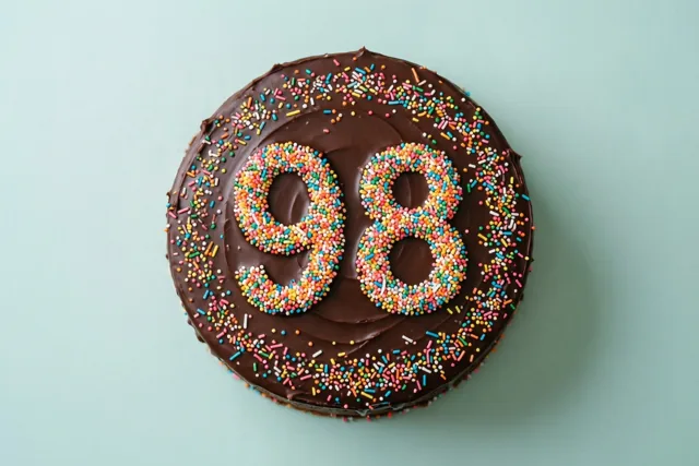 Van bovenaf is een ronde chocoladetaart met chocoladeglazuur, gekleurde suikerstrooisels en het getal 98 te zien tegen een lichtblauwe achtergrond.