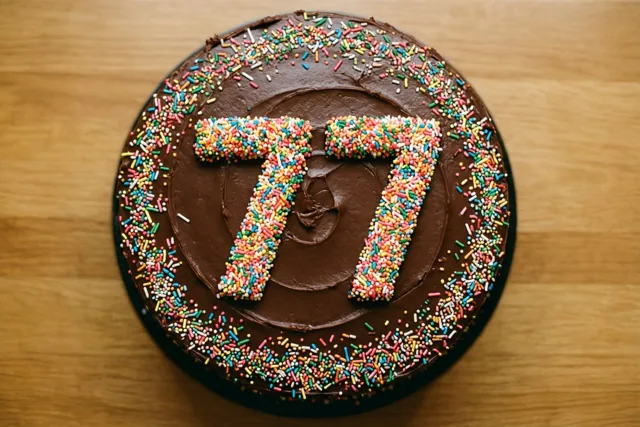Een bovenaanzicht toont een ronde chocoladetaart met chocoladeglazuur en 77 van kleurrijke suikerstrooisels op een houten ondergrond.