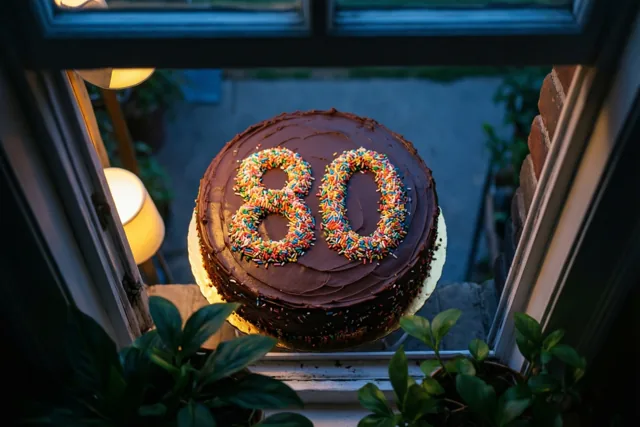 Van bovenaf is een ronde chocoladetaart met chocoladeglazuur en het getal 80 van gekleurde suikerstrooisels te zien op een vensterbank, met een raam eromheen en groene planten op de voorgrond in warm binnenlicht.