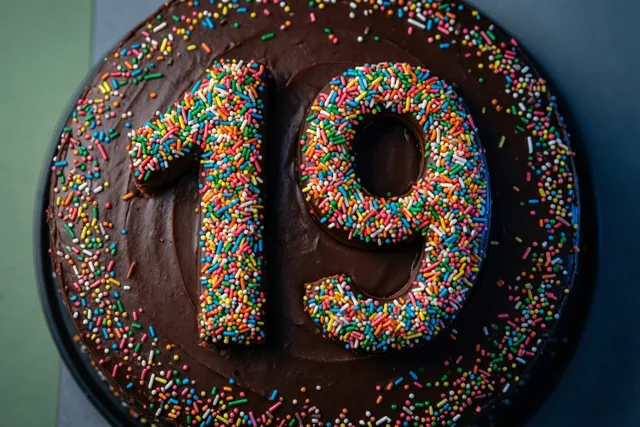 Van bovenaf is een ronde chocoladetaart op een donker bord te zien, met 19 van kleurrijke suikerstrooisels en losse strooisels erop.