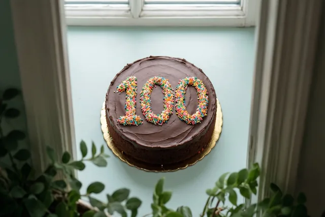 Een ronde chocoladetaart met 100 in gekleurde suikerstrooisels staat op een gouden taartbodem op een lichtblauw oppervlak voor een wit raamkozijn, met groen op de voorgrond.