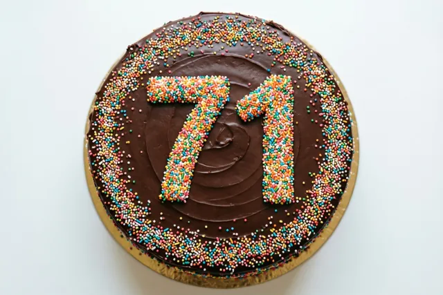 Een ronde chocoladetaart met glazuur en suikerstrooisels die 71 vormen staat op een gouden kartonnen taartbodem tegen een witte achtergrond.
