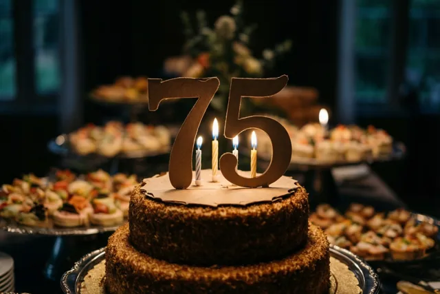 Een chocoladetaart met meerdere verdiepingen, een papieren 75 en brandende kaarsjes staat op een zilveren schaal op een buffet met wazige schalen eten op de achtergrond.
