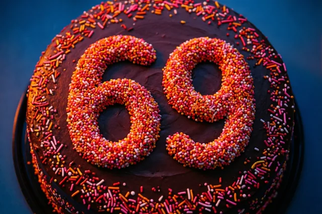 Van bovenaf is een ronde chocoladetaart te zien met het getal 69 in kleurrijke suikerstrooisels tegen een donkerblauwe achtergrond.
