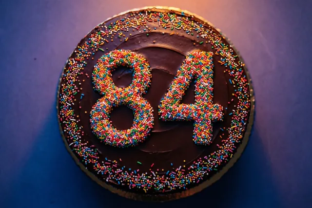Een ronde chocoladetaart met 84 van gekleurde suikerstrooisels is van bovenaf gefotografeerd tegen een donkerblauwe achtergrond in warm licht.