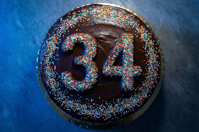 Een ronde chocoladetaart met het getal 34 van gekleurde suikerstrooisels is van bovenaf te zien tegen een donkerblauwe achtergrond met structuur.
