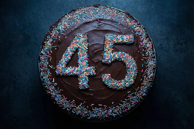 Een ronde chocoladetaart met 45 van gekleurde suikerstrooisels is van bovenaf te zien tegen een donkere achtergrond met structuur.