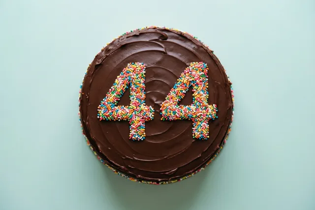 Een ronde chocoladetaart met glad glazuur, 44 in kleurrijke suikerstrooisels en strooisels langs de rand is van bovenaf te zien tegen een lichtblauwe achtergrond.