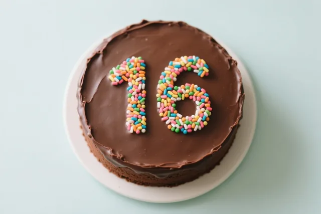 Van bovenaf is een ronde chocoladetaart met 16 in gekleurde suikerstrooisels te zien, op een wit bord tegen een lichtblauwe achtergrond.