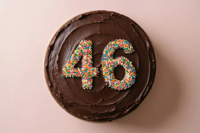 Van bovenaf is een ronde chocoladetaart te zien met 46 van gekleurde suikerstrooisels tegen een effen lichtroze achtergrond.