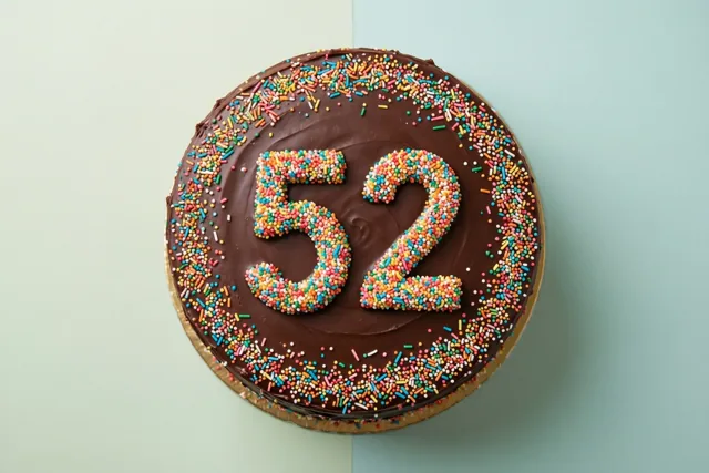 Een ronde chocoladetaart met 52 van gekleurde suikerstrooisels is van bovenaf te zien tegen een pastelgroene en pastelblauwe achtergrond.