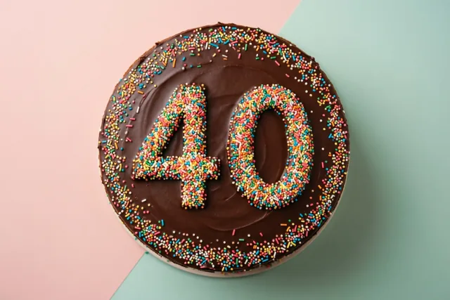 Een ronde chocoladetaart met 40 van gekleurde suikerstrooisels is van bovenaf te zien tegen een achtergrond met een roze en een blauwgroen deel.