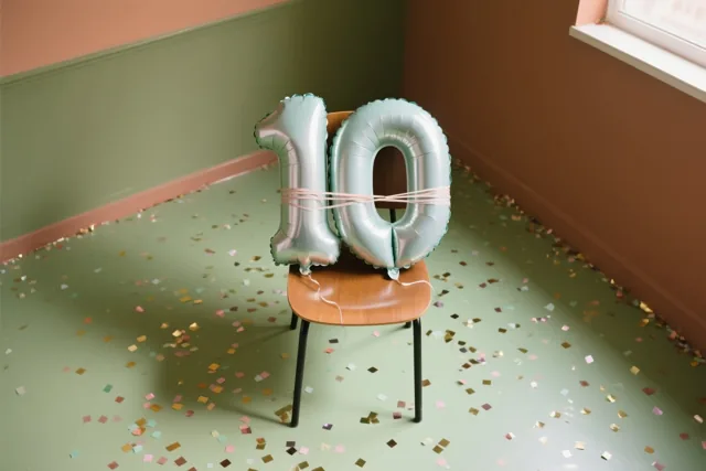 Zilver- en pastelkleurige cijferballonnen die samen 10 vormen zijn vastgebonden aan een houten stoel, met confetti op de vloer en groen- en terracottakleurige muren.
