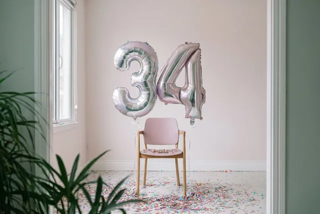 Zilveren en pastelkleurige cijferballonnen vormen 34 aan een roze stoel in een lichte kamer met confetti, roze muren en groene planten, gezien door een deuropening.