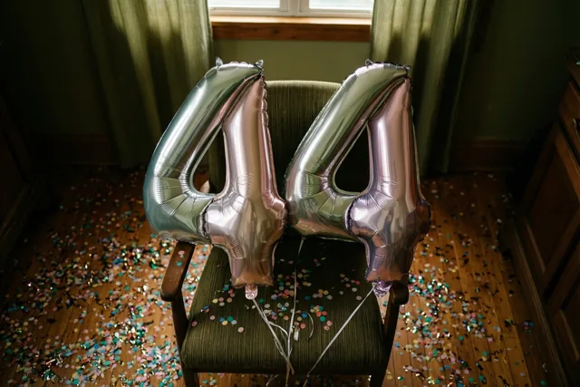 Twee grote zilver- en pastelkleurige ballonnen vormen 44 aan een groen beklede stoel op een houten vloer met kleurrijke confetti, verlicht door een raam.